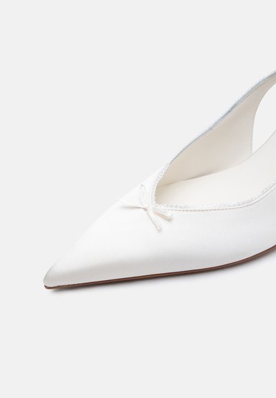 Chaussure plate blanche à bout pointu avec un petit nœud décoratif et une bordure festonnée sur le bord, photographiée sur un fond clair.