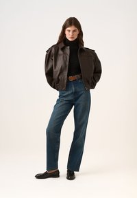 Hnědá kožená bomber bunda s žebrovaným lemem, černý rolák, modré džíny s vysokým pasem a černé mokasíny. Neutrální pozadí.