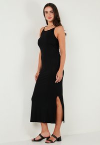 Black sleeveless maxi dress with side slits, fitted design, smooth texture, and ankle-length; paired with black strappy sandals.