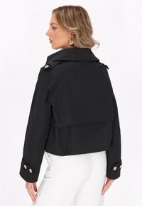 Giacca cropped nera con ampio colletto, polsini con bottoni e parte posteriore a strati. Realizzata in materiale liscio, con dettagli in metallo argentato.