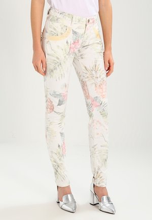 Kvinde iført slim-fit hvide jeans med pastelfarvede tropiske blomster- og ananasmønstre, kombineret med sølvfarvede højhælede sko.