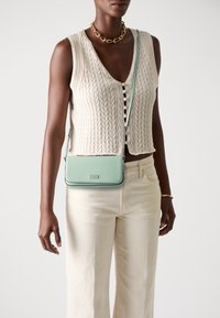 Mint green crossbody bag with a smooth leather finish, silver hardware, and a chain strap, worn over a knitted beige vest and cream pants.