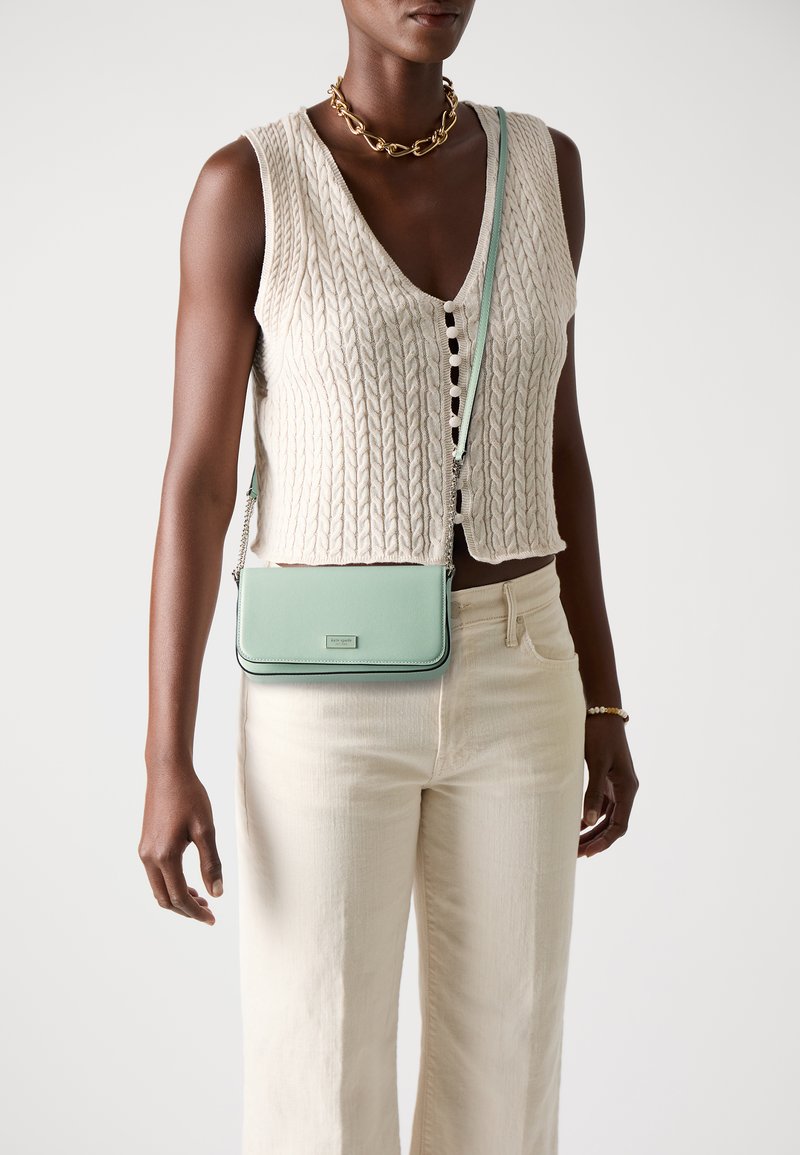 Mint green crossbody bag with a smooth leather finish, silver hardware, and a chain strap, worn over a knitted beige vest and cream pants.
