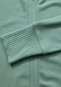 Pull en laine vert menthe avec un col en V, des poignets côtelés et une texture lisse. Inclut des coutures verticales sur le devant.