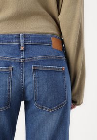 Jeanși din denim de culoare albastru închis, cu o textură netedă, având o etichetă din piele maro și două buzunare spate cu detalii de cusătură contrastante.