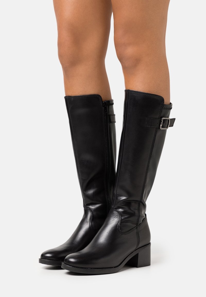 Jambes chaussées de bottes hautes en cuir noir, avec une fermeture éclair sur le côté et de petits talons carrés, posées devant un fond clair uni.