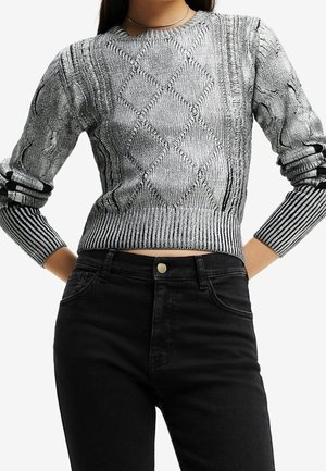 Vrouw draagt een nauwsluitende grijze kabelgebreide trui met diamantpatronen en zwarte hoge taille jeans, handen in de zij.