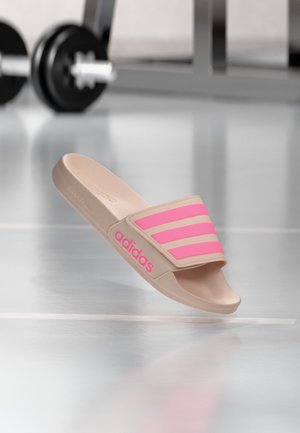 Μπεζ σαγιονάρα με ροζ ρίγες και λογότυπο adidas, αιωρείται πάνω από το πάτωμα του γυμναστηρίου με θολό αλτήρα στο παρασκήνιο.