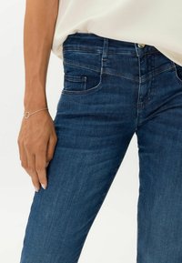 Jeans de mezclilla azul oscuro con una textura suave, que presentan un diseño clásico de cinco bolsillos y detalles de costura sutiles. Hardware de botón plateado visible.