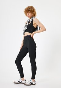 Zwarte cropped sporttop gecombineerd met hoge zwarte leggings. Een zwart-wit gestreepte sjaal nonchalant om de schouders gedrapeerd.