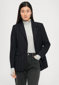 Tummansininen pinstripe-puku, jossa on muotolattu istuvuus, kaksi etutaskua, yksittäinen nappisulku ja tekstuurinen kangas harmaan poolopaidan päällä.