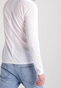 Person wearing a white long-sleeve shirt and blue jeans, visible from the back and side with a light gray background.