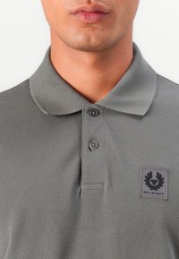 Graues Polohemd aus strukturiertem Stoff, mit drei Knöpfen und einem gestickten Logo-Patch auf der linken Brust. Kurzes Kragen-Design.
