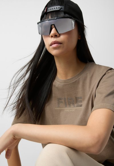 Brown T-shirt with "FIRE" in rhinestones, paired with large gray sunglasses and a black cap. Smooth fabric; hair flows around the face.
