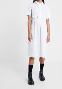 Abito camicia bianco a maniche corte con bottoni frontali e vita arricciata, dotato di colletto e orlo fino al ginocchio. Indossato con stivaletti neri e calze.