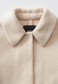 Beige wollen jas met een zachte textuur, voorzien van een klassieke kraag en een sluiting aan de voorkant, met een zwart merketiket op de hals.