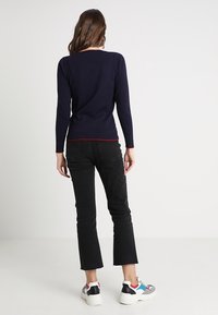 Donna in piedi, indossa una maglia a maniche lunghe blu navy, jeans neri cropped e sneakers bianche con dettagli rossi, blu e grigi, su uno sfondo semplice.