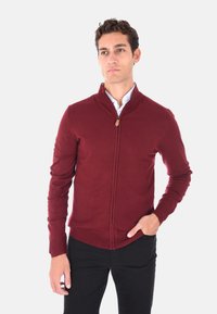 Maglione borgogna con zip, caratterizzato da un colletto alto, polsini e orlo a coste. Realizzato in morbido materiale a maglia, con un dettaglio in pelle marrone per la zip.