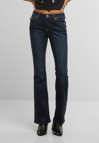 Jeans a zampa d'elefante blu scuro con vita media, caratterizzati da cuciture arancioni, tasche frontali e chiusura con bottone argentato. Indossati con stivali neri.