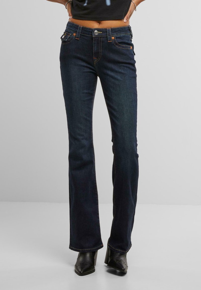 Jeans a zampa d'elefante blu scuro con vita media, caratterizzati da cuciture arancioni, tasche frontali e chiusura con bottone argentato. Indossati con stivali neri.