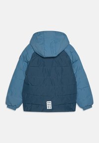 Jachetă captusită colorată în nuanțe de albastru, cu glugă, manșete elastice, cusături orizontale și o patch reflectorizantă albă în partea de jos a spatelui.