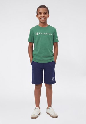 Maglietta di cotone verde con logo "Champion", abbinata a pantaloncini blu navy e sneakers beige. Vestibilità casual con maniche corte e scollo rotondo.
