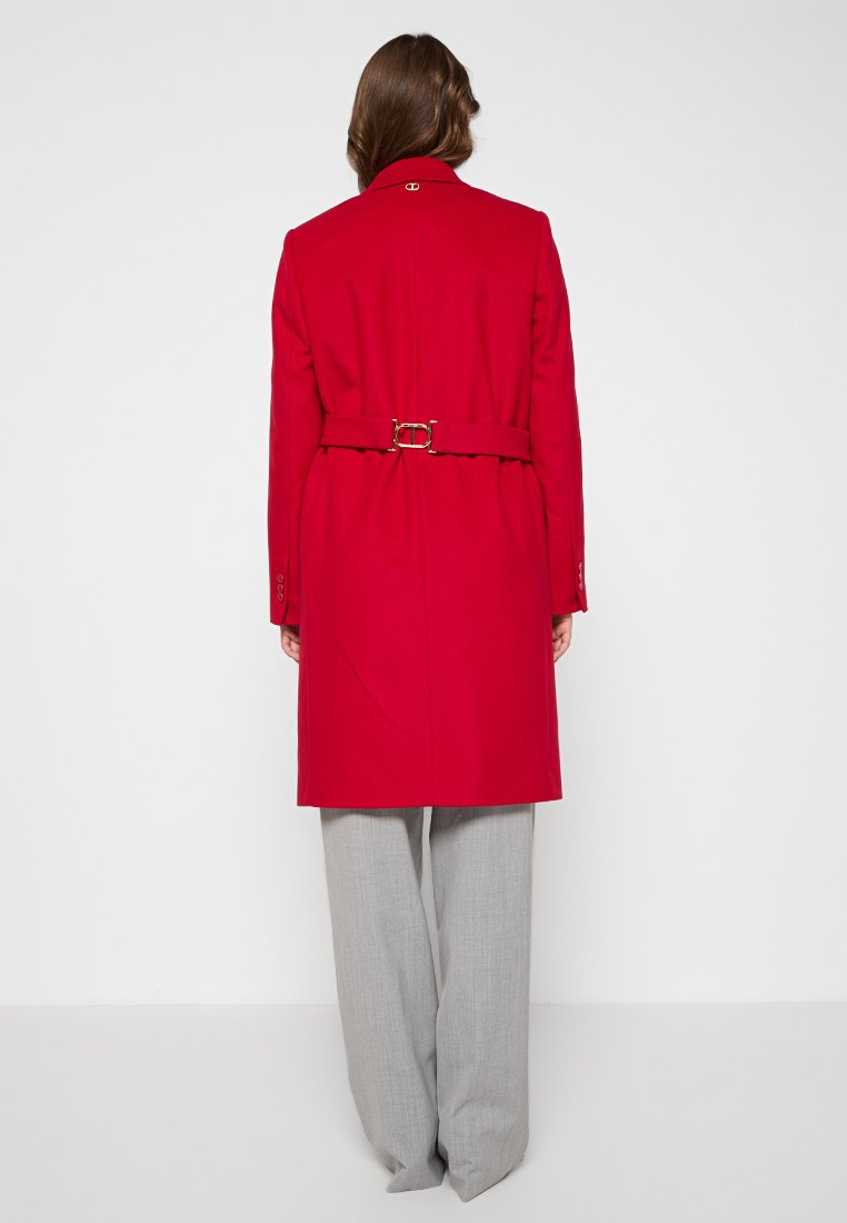 Cappotto in misto lana rosso con vita cintata, dotato di un colletto classico e dettagli con bottoni. Abbinato a pantaloni a gamba larga grigio chiaro.