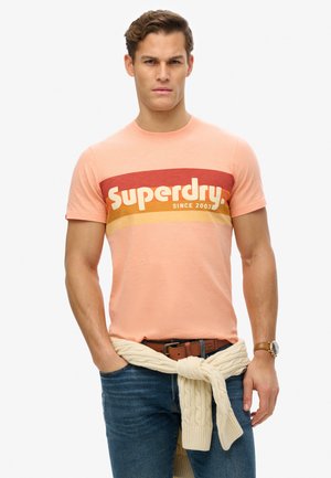 Short-sleeve peach t-shirt with a bold, multicolored "Superdry" logo and stripes. Worn with dark denim and a light knit sweater tied at the waist.