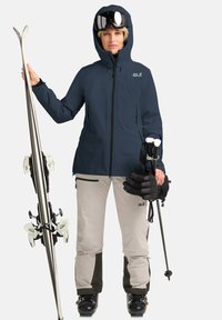 Skiausrüstung bestehend aus einer navyblauen wasserdichten Jacke, beigen Skihosen mit schwarzen Akzenten, schwarzen Handschuhen, Skistöcken und Skiern mit Bindungen.