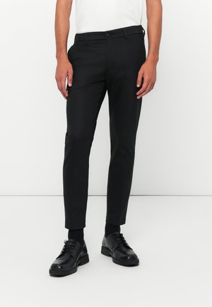 Zwarte broek met een slim fit ontwerp, gemaakt van soepel stof. Beschikt over een tailleband met riemlussen en zijzakken. Draagt men met zwarte schoenen.