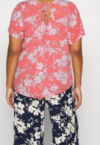 Haut à manches courtes floral en corail avec un dos en trou de serrure noué. Associez-le à un pantalon ample bleu marine avec un motif floral blanc.