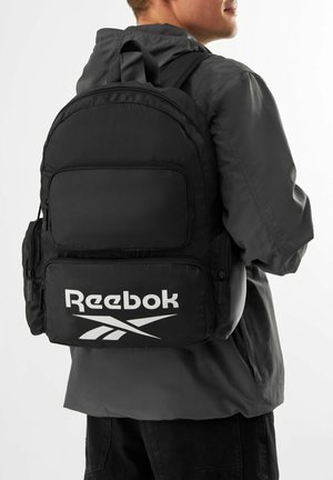 Mężczyzna w ciemnej kurtce z czarnym plecakiem Reebok z wieloma zapinanymi na suwak kieszeniami, przedstawiony z tyłu i z boku.