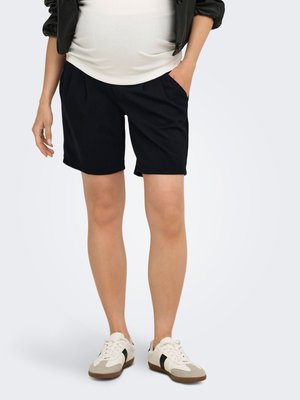 Person som har på seg svarte shorts, hvite joggesko med svarte striper, en hvit topp og en svart jakke, står med en hånd i lommen.