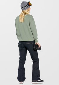 Groene sweatshirt en zwarte skibroek met zijritsen. Zwarte beanie met streep patroon en skibril. Zwart sneakers met witte zool.