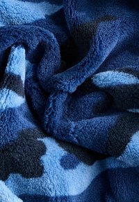 Minkštas antklodė skirtingų mėlynos spalvos atspalvių, su kamuflažo raštu. Švelni, tekstūruota medžiaga su puriu, jaukiu išvaizdu.