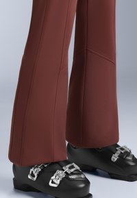 Hnedé lyžiarske nohavice s rozšírenými nohavicami, s plochými švami a hladkým povrchom, spárované s čiernymi lyžiarskymi topánkami so striebornými prackami.