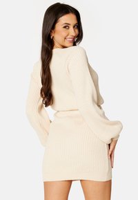 Abito a maglia a coste color crema, con maniche lunghe, vita aderente e lavorazione in tessuto testurizzato. Presenta un colletto rotondo e lunghezza fino a metà coscia.