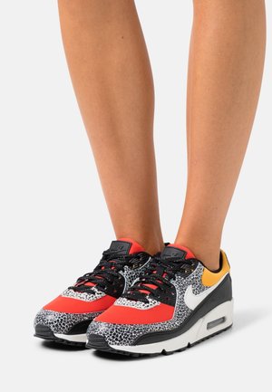 Buty sportowe w czarno-biało-czerwony wzór, z teksturowaną cholewką, żółtym akcentem i widoczną jednostką amortyzującą powietrze w podeszwie.