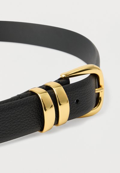 Črni usnjeni pas z teksturirano površino, ki ima sijoč zlati zaponko in dva ujemajoča zlata zanka na belem ozadju.