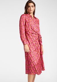 Vestido camiseros rosa con un patrón de cadena geométrica en naranja y malva. Presenta mangas largas, un cinturón a la cintura y un cuello de camisa. Tela suave.