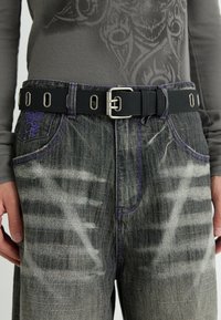 Donkerdenim jeans met vervaagde en versleten gebieden, geaccentueerd met paarse stiksels en een zwarte riem met een metalen oogjes.