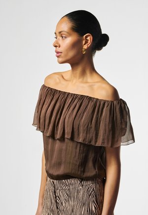 Frau mit dunklen Haaren im Dutt trägt eine braune schulterfreie Bluse und Zebrahose, schaut nach links vor einem schlichten weißen Hintergrund.