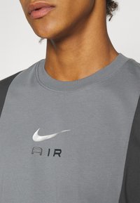Šedé tričko s krátkým rukávem a kontrastními tmavými rukávy, s stříbrným logem Nike swoosh a textem "AIR" v černé barvě. Měkká textilní struktura.