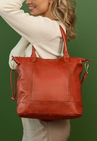 Leren draagtas in burnt orange, met dubbele handvatten en een afneembare schouderband. Gladde textuur met een elegante, gestructureerde vorm.