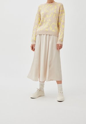 Persona con un suéter beige y amarillo con estampado, falda midi color crema, calcetines blancos y zapatillas blancas, de pie frente a un fondo liso.