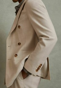 Jachetă de costum dublu din beige deschis, cu butoane din lemn, textură subtilă și rever cu crestătură. Buzunarul lateral este vizibil, însoțit de pantaloni asortați.