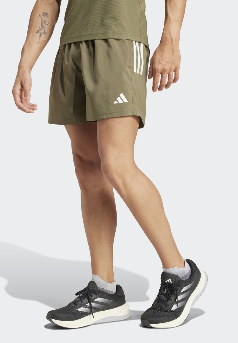 Olive green athletic shorts with white striped accents and logo; worn with black running shoes featuring white stripes and grey socks.