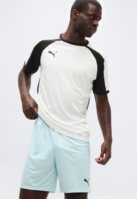 Αθλητικός άνδρας που ρυθμίζει ανοιχτό μπλε σορτς Puma, φοράει λευκό και μαύρο αθλητικό μπλουζάκι Puma, στέκεται μπροστά σε απλό ανοιχτό φόντο.