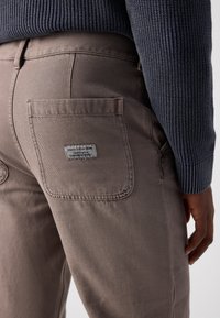 Pantalons en coton beige avec une poche arrière dotée d'une étiquette tissée. Le matériau présente une légère texture et une coupe classique.