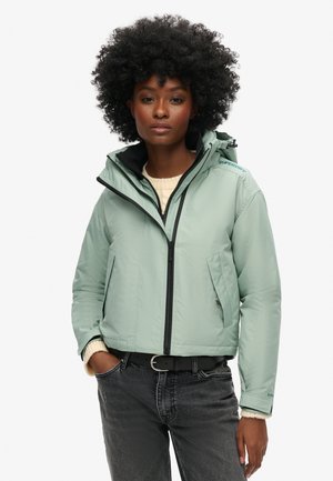 Light green puffer jacket with a zippered front, adjustable hood, and two diagonal side pockets, layered over a cream sweater and black belt.
